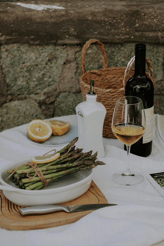 Ein Tisch mit frischem Spargel, einem Glas Weißwein, Olivenöl, Zitronenscheiben und einer Flasche Wein, vor einer Steinmauer.
